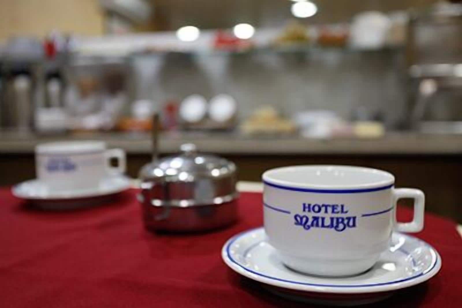 Un entorno de hotel con una taza y platillo con la etiqueta "HOTEL MALIBU" y una tetera sobre un mantel rojo.
