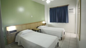 Habitación de hotel con dos camas individuales, un ventilador de techo, una mesita de noche con lámpara, una ventana con cortina y un aire acondicionado instalado en la pared.