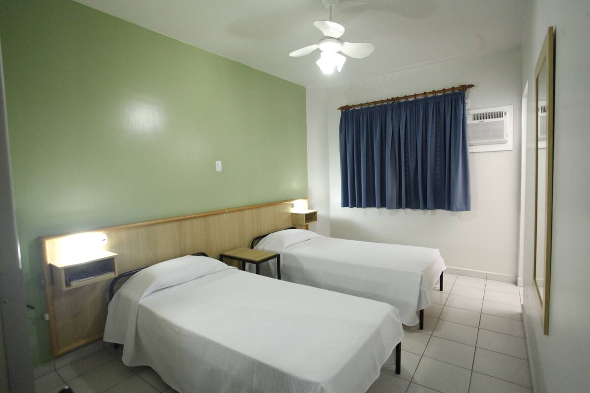 Habitación de hotel con dos camas individuales, un ventilador de techo, una mesita de noche con lámpara, una ventana con cortina y un aire acondicionado instalado en la pared.