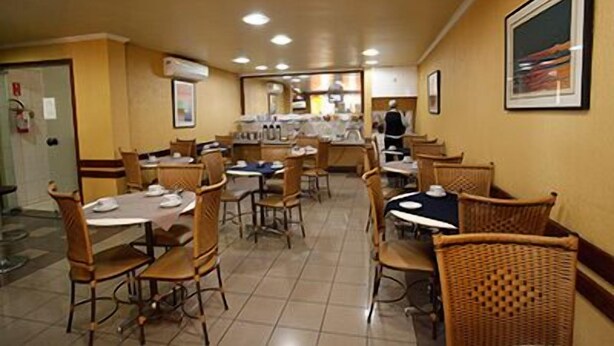 Comedor de hotel con varias mesas y sillas, una cocina al fondo y un cuadro en la pared.