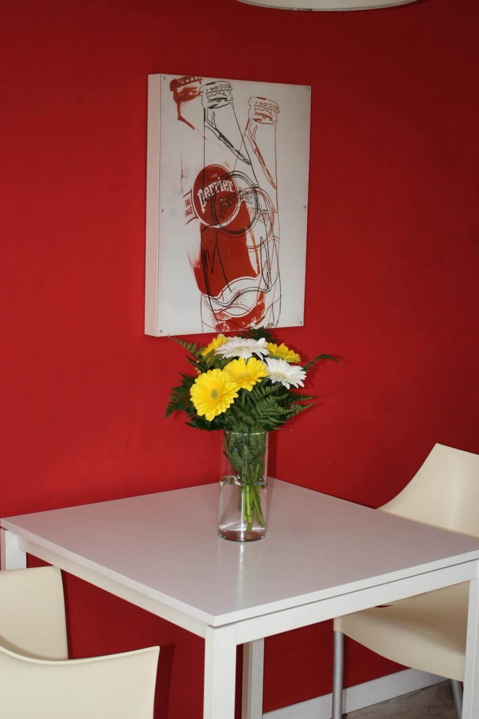 A white table with a vase of yellow and white flowers, a red wall, and a poster with abstract designs.