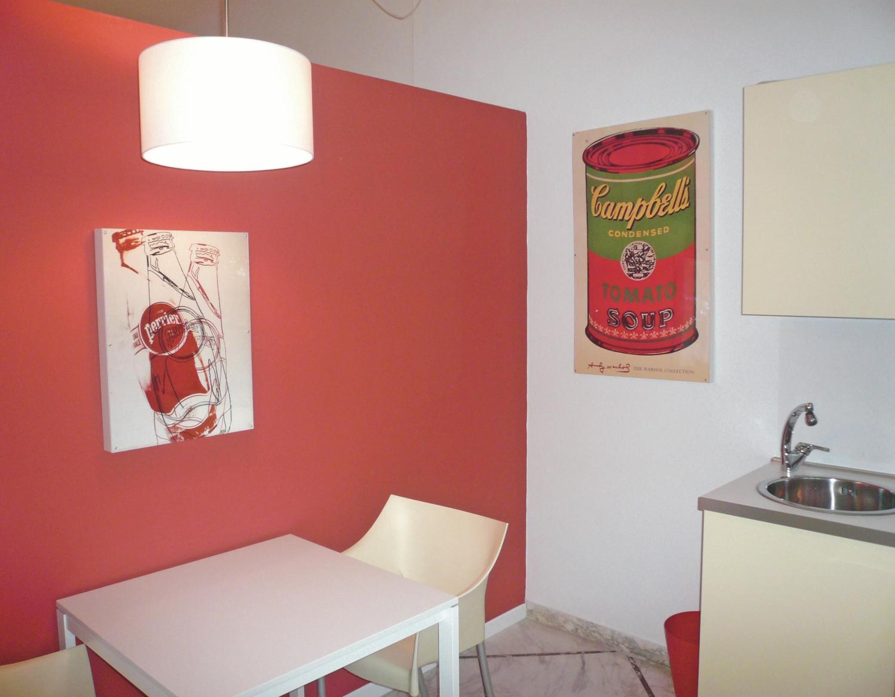 A room with a red wall, a white table, a white chair, a hanging lamp, a poster of Campbell's Tomato Soup, and a sink.