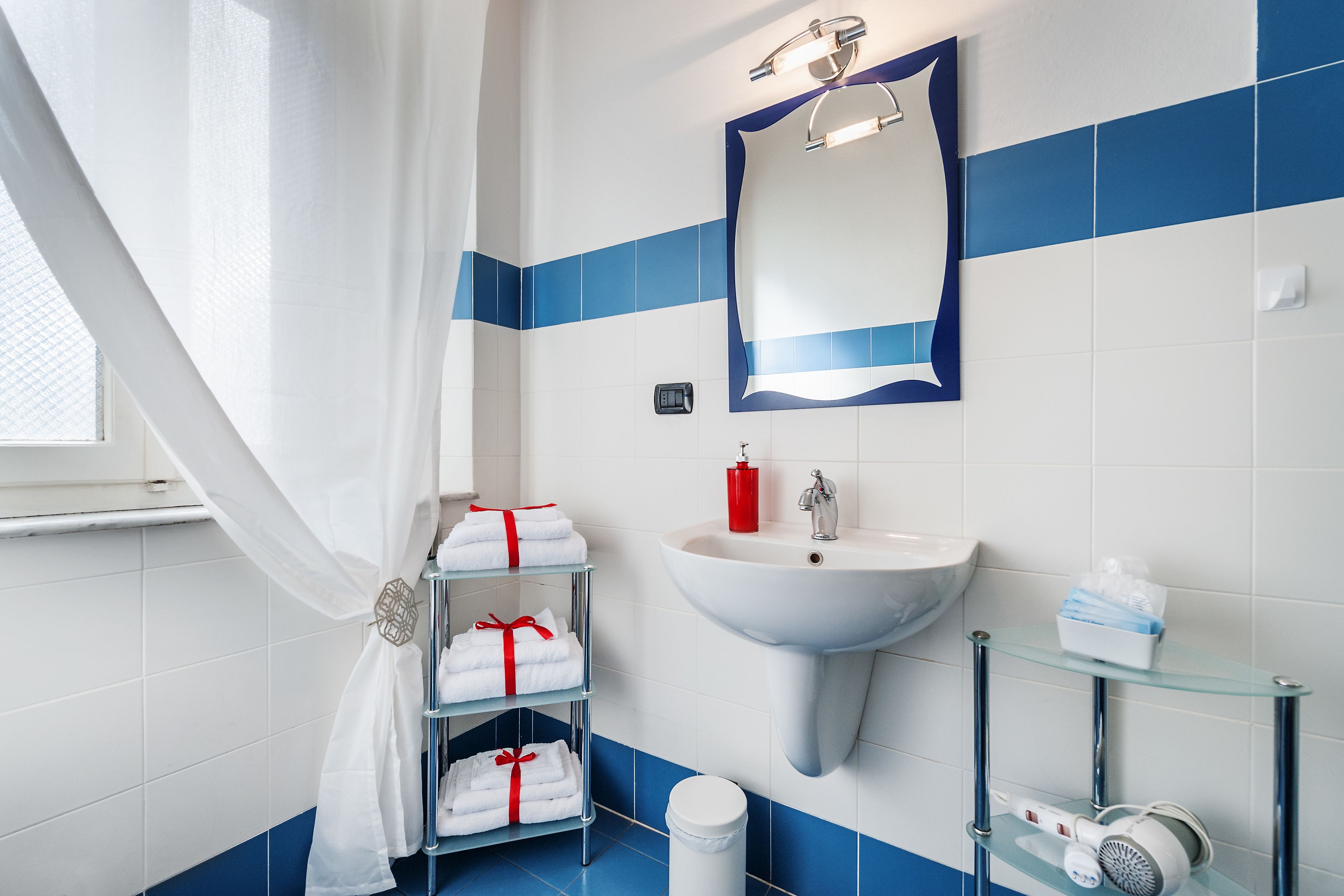 Un baño con lavamanos blanco, azulejos azules, un estante de vidrio con artículos de aseo personal y un perchero con toallas dobladas con cuidado.