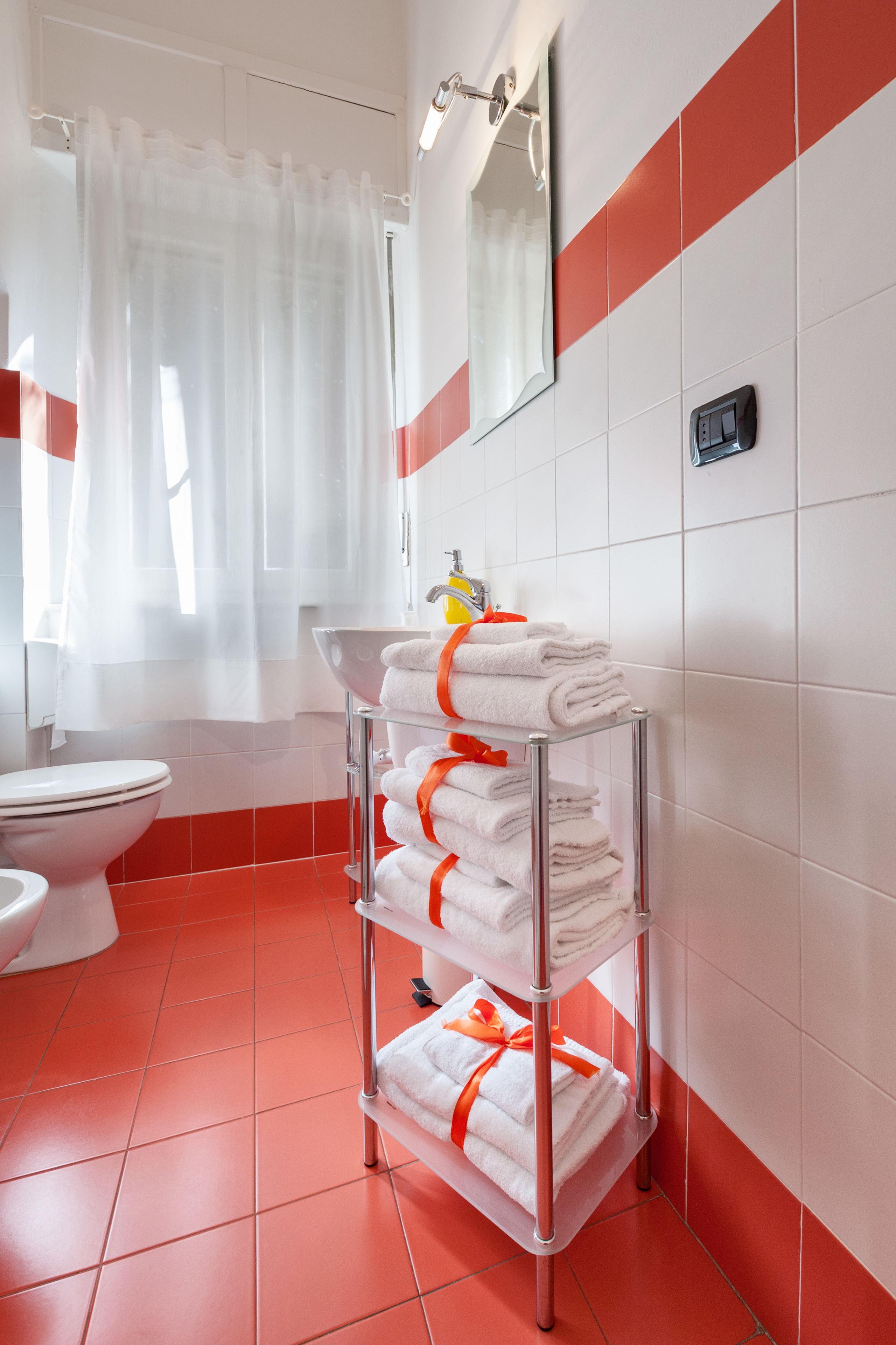 Ein Badezimmer mit weiß-roten Fliesen, einer weißen Toilette, einem Waschbecken mit Spiegel und einem Handtuchhalter mit gefalteten Handtüchern.