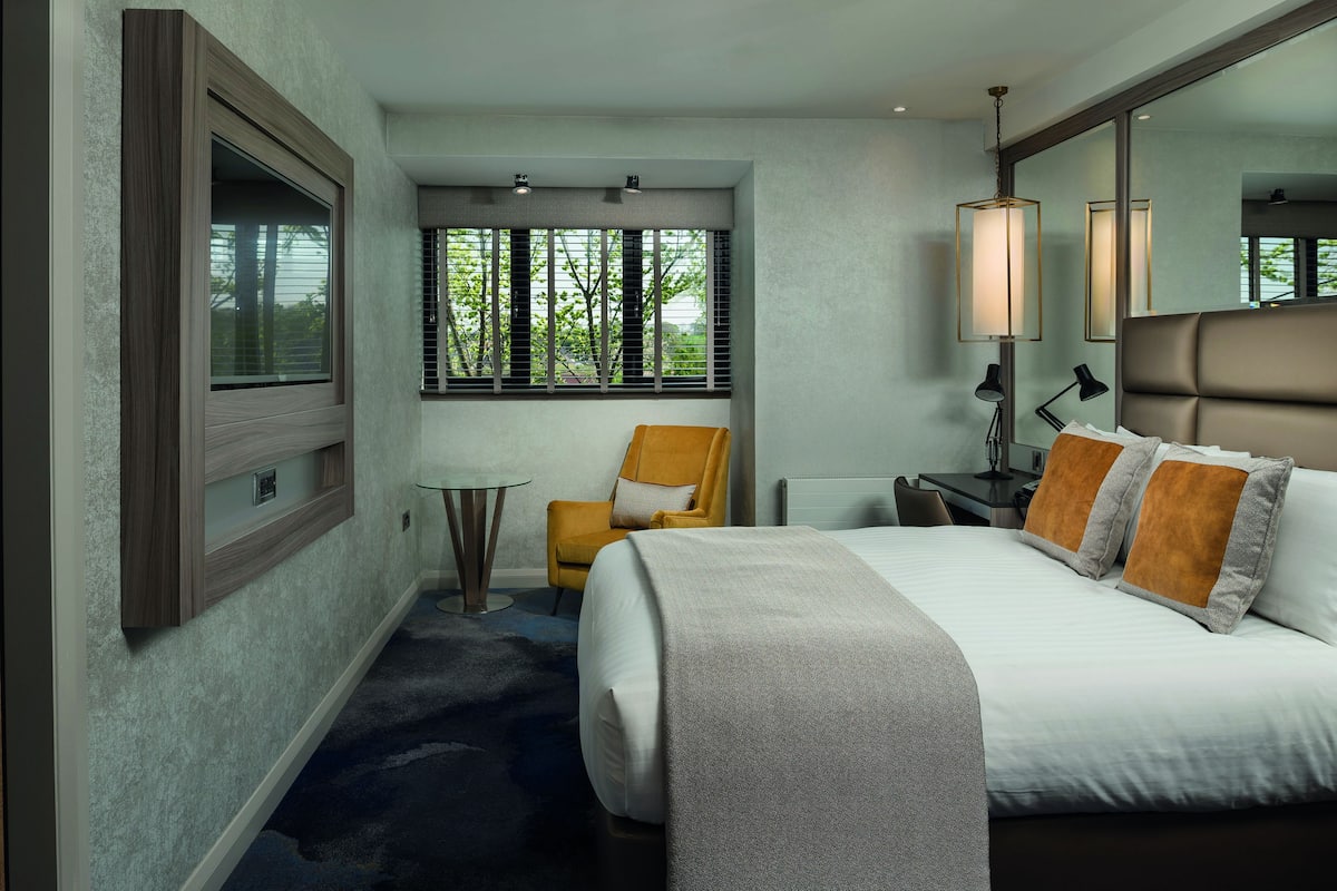 A hotel room with a bed, a chair, a desk, and a window with blinds.