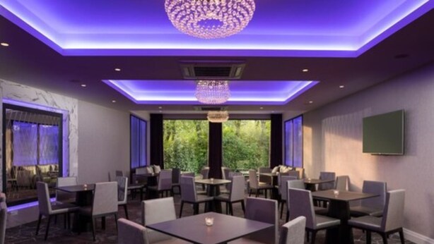 Un restaurante con varias mesas y sillas, un candelabro y ventanas amplias con vistas a la vegetación.