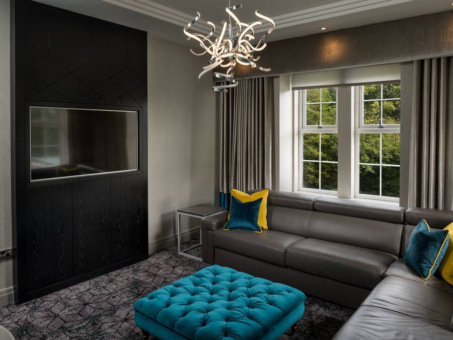 A modern living room with a dark wood panel wall, a large flat-screen TV, a grey leather sofa with blue and yellow cushions, a teal tufted ottoman, and a unique chandelier.