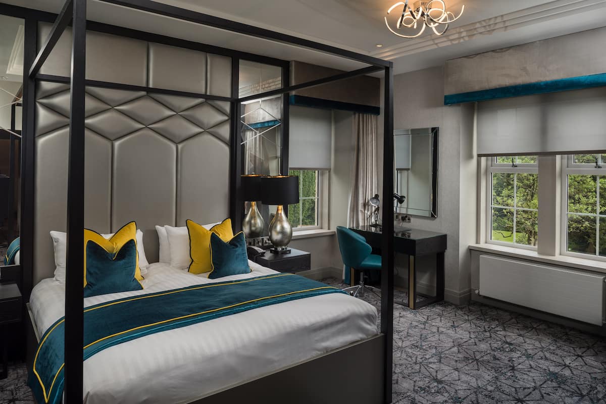 A four-poster bed with a modern design, a desk with a chair, and a large window with a view of greenery.