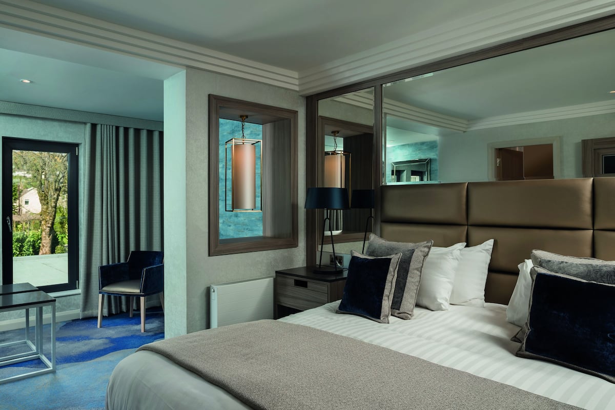 A modern hotel room with a large bed, a desk, and a chair. There is a window with curtains, a mirror, and a lamp on the nightstand.