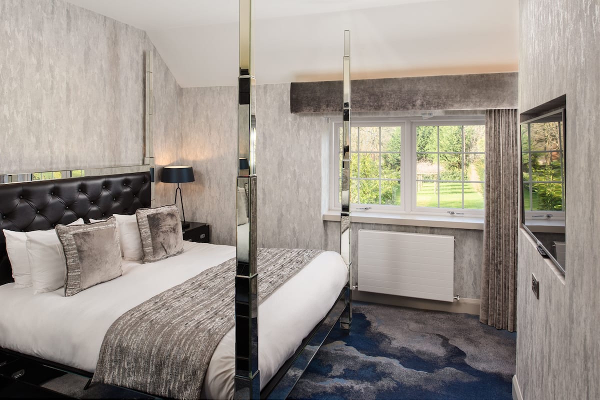 A modern hotel room with a large bed, a window with a view of greenery, a radiator, and a tall floor lamp.