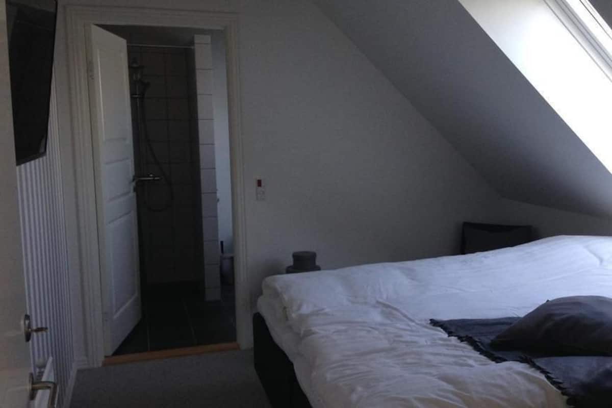 Un dormitorio con techo inclinado, una cama, una mesita de noche y una puerta que da a un baño.