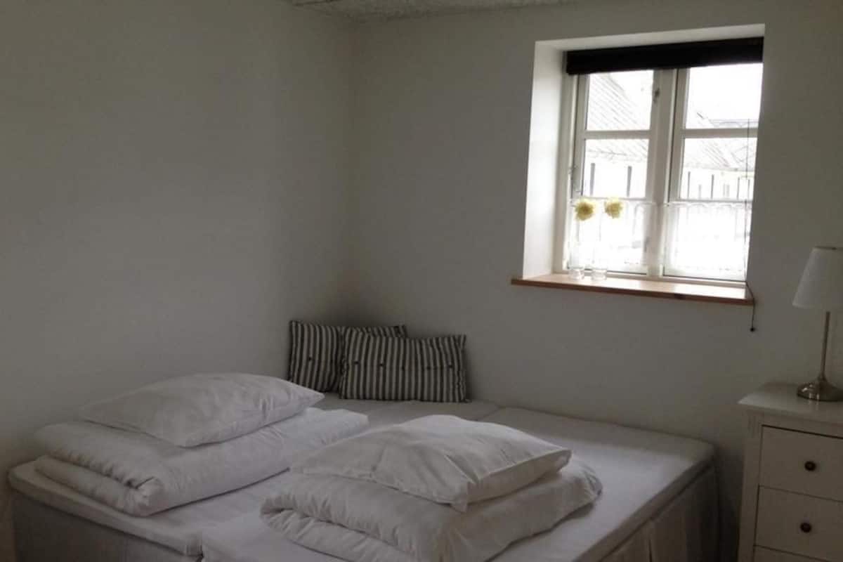 Un dormitorio con cama, ventana, mesita de noche y un velador.