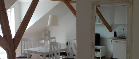 A dining area with white chairs and a table, a kitchen with white cabinets, and a wooden beam structure.