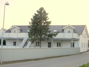 Un edificio de dos pisos con fachada blanca, una escalera que lleva al nivel superior y un árbol en la parte delantera.