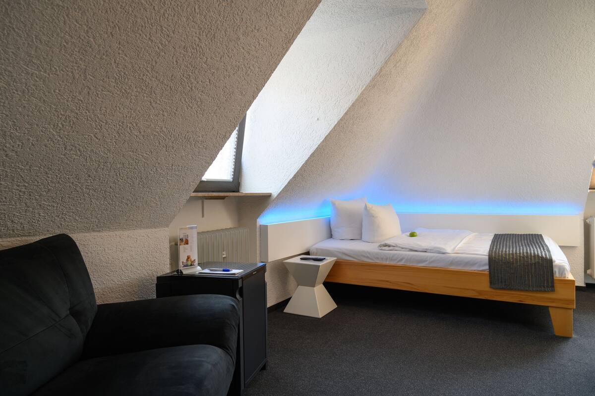 Ein modernes Schlafzimmer mit schrägen Wänden, einem hölzernen Bett, einem schwarzen Ledersessel, einem kleinen Nachttisch und einem Beistelltisch.