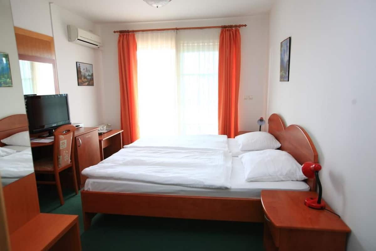 A hotel room with a single bed, a desk with a computer, a television, and a red lamp.