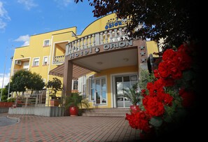 L’entrée d’un hôtel avec une façade jaune, un balcon et des fleurs rouges au premier plan.