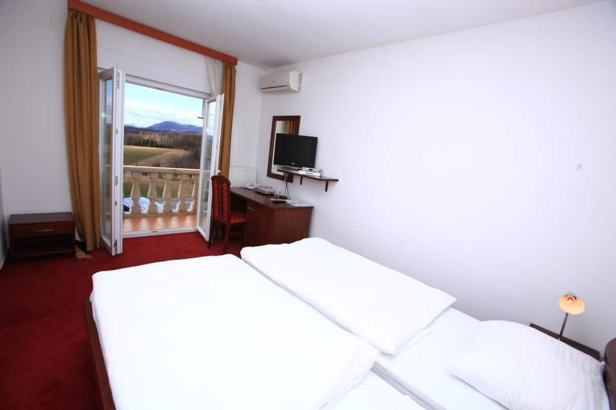 A hotel room with two beds, a desk with a TV, a window with a view of the countryside, and an air conditioning unit.