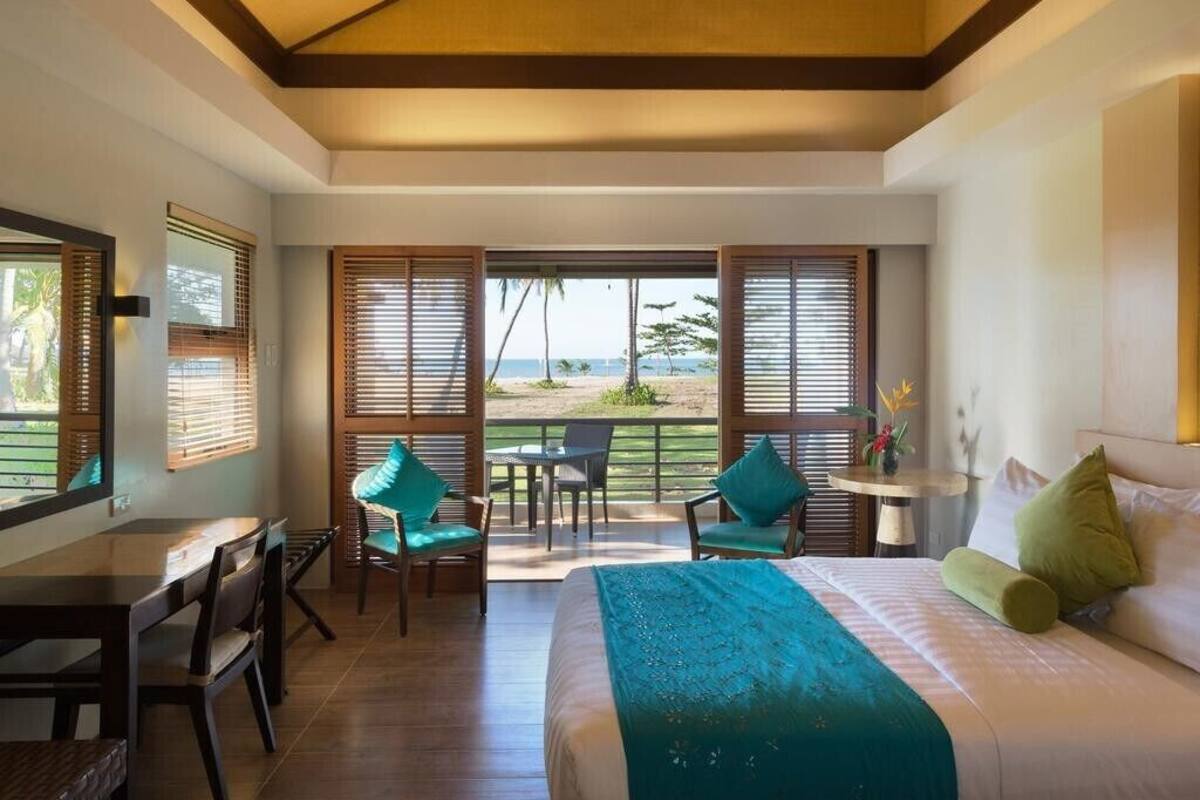 A hotel room with a bed, a desk, two chairs, a small table, and a view of the beach.