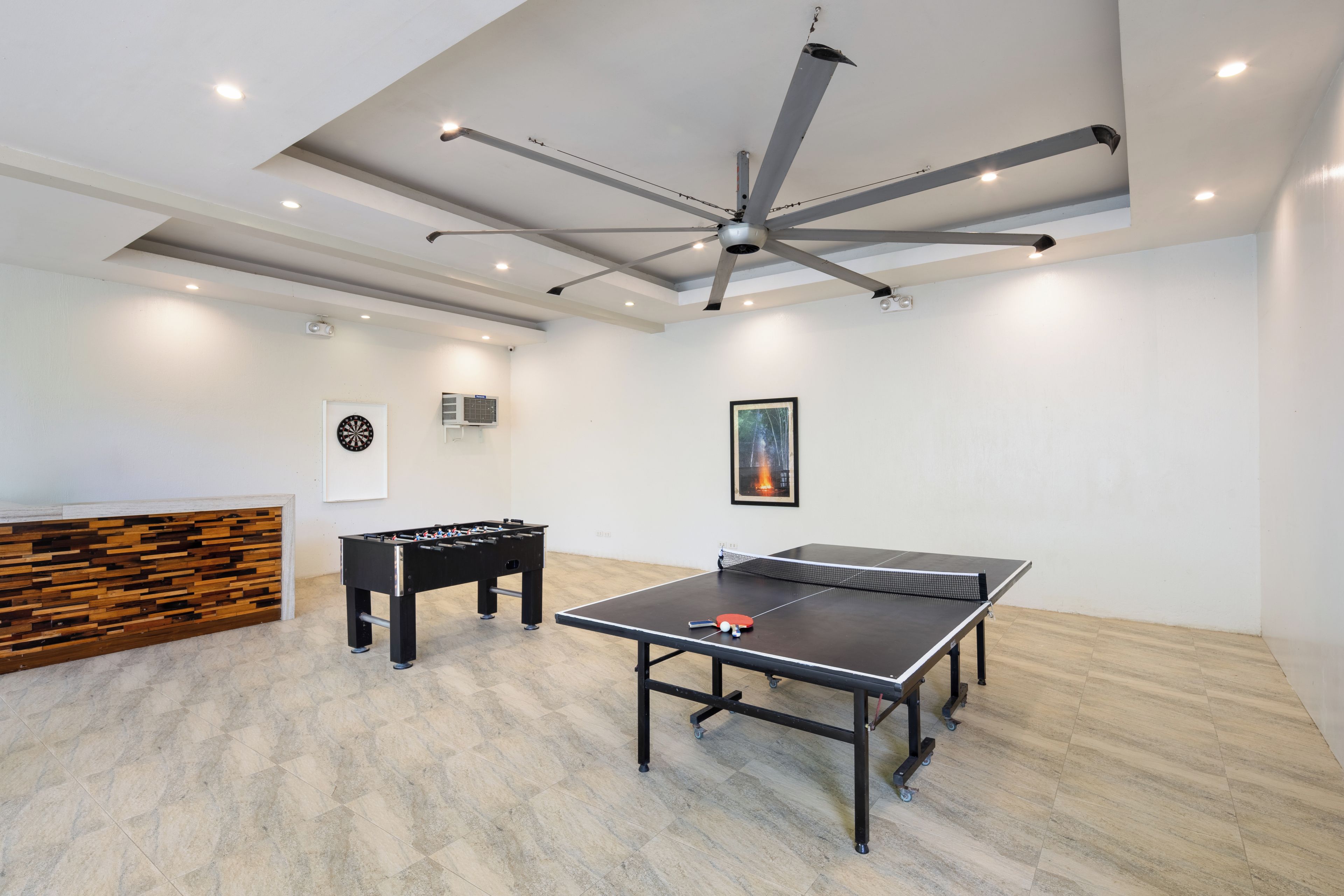 A room with a ping pong table, foosball table, and a ceiling fan.