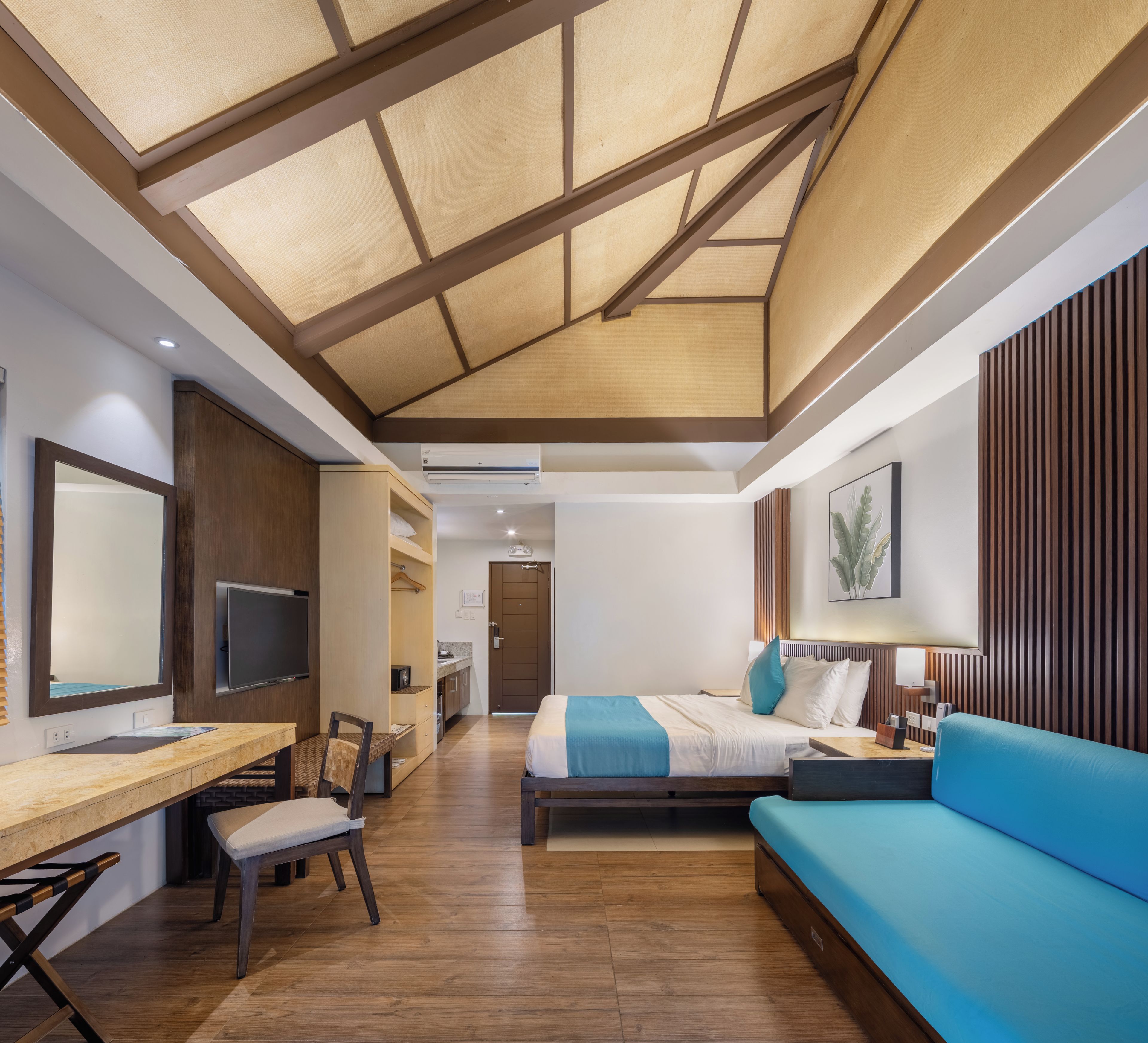 Una habitación de hotel moderna con suelo de madera, cama con ropa de cama azul, un escritorio con silla, un televisor y un ventanal grande.