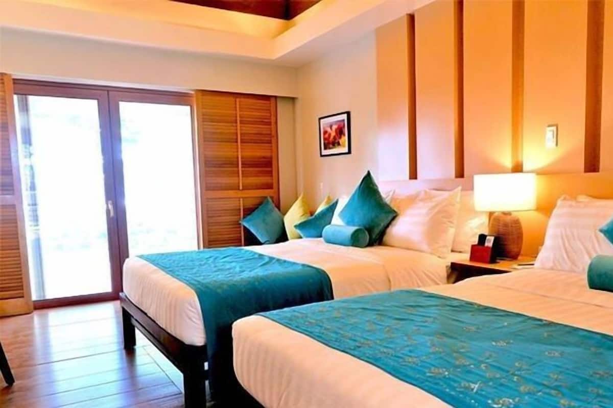 A hotel room with two beds, a wooden wardrobe, a picture on the wall, and a nightstand with a lamp.