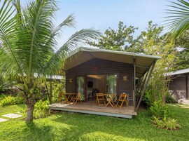 Una pequeña cabaña de madera con un porche cubierto, mobiliario exterior y un jardín con palmeras.