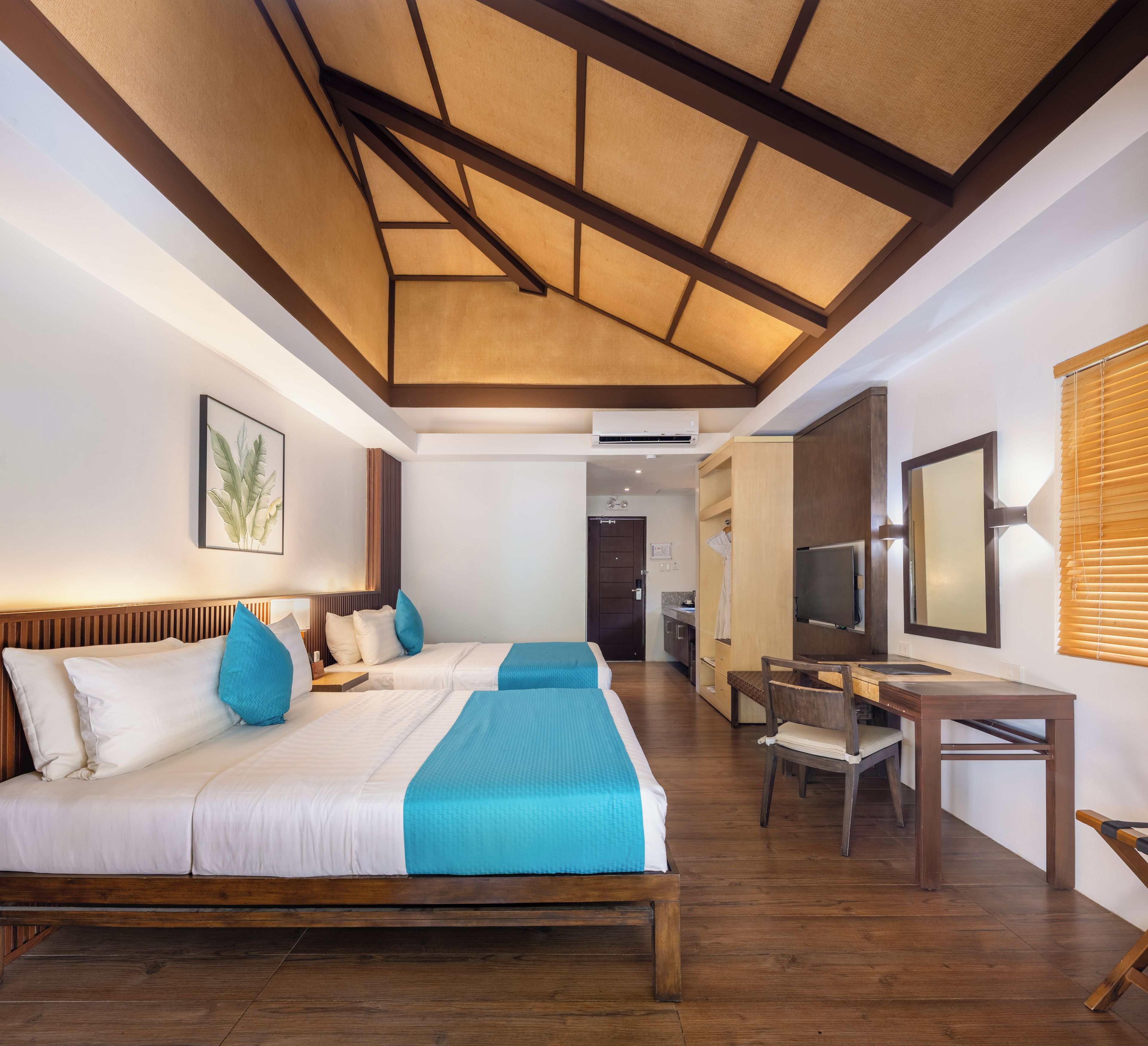 A hotel room with a wooden ceiling, a bed with white and blue bedding, a desk with a chair, and a television.