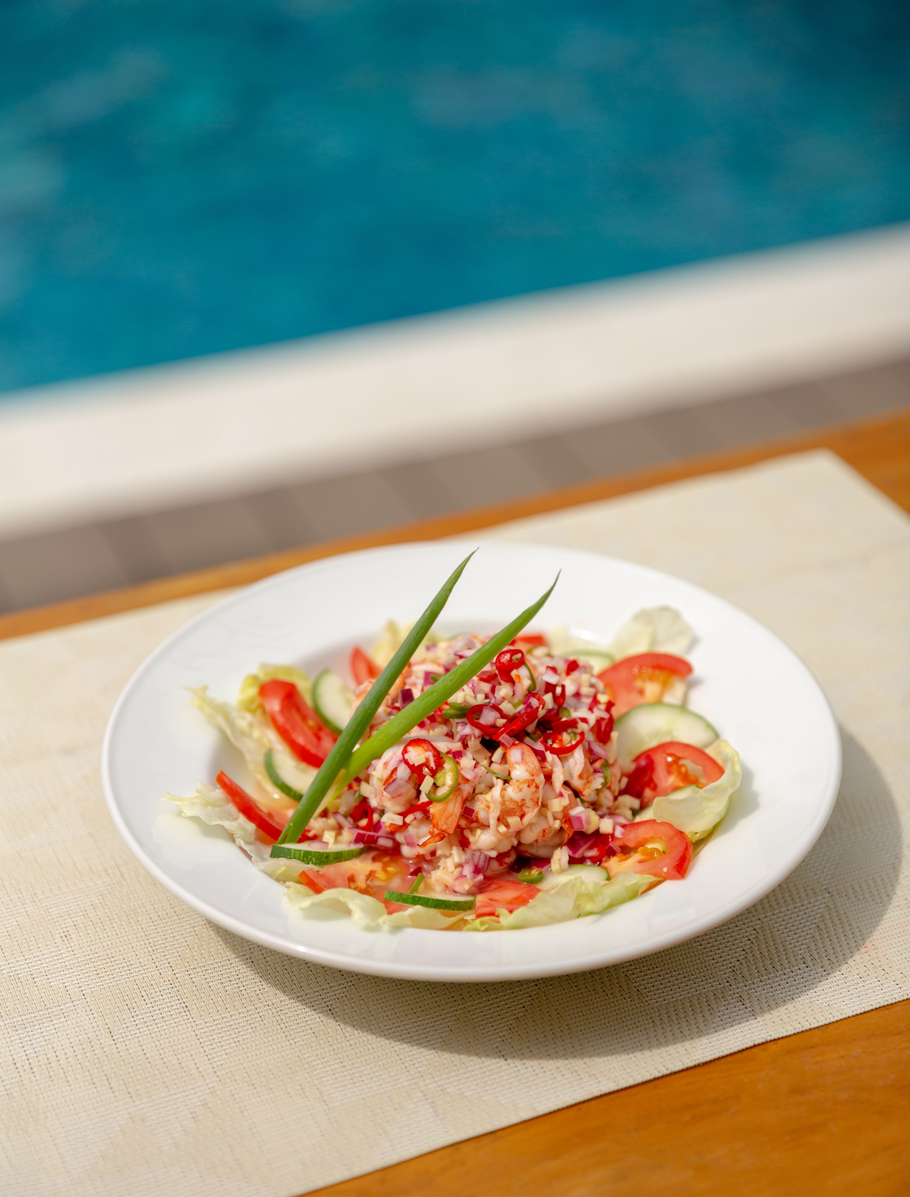 A plate of shrimp salad with vegetables and green onions, placed on a table by a poolside.