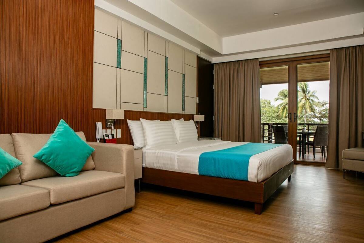 A hotel room with a wooden floor, a bed with white linens and a blue accent, a beige sofa with teal pillows, and a large window with a view of palm trees.