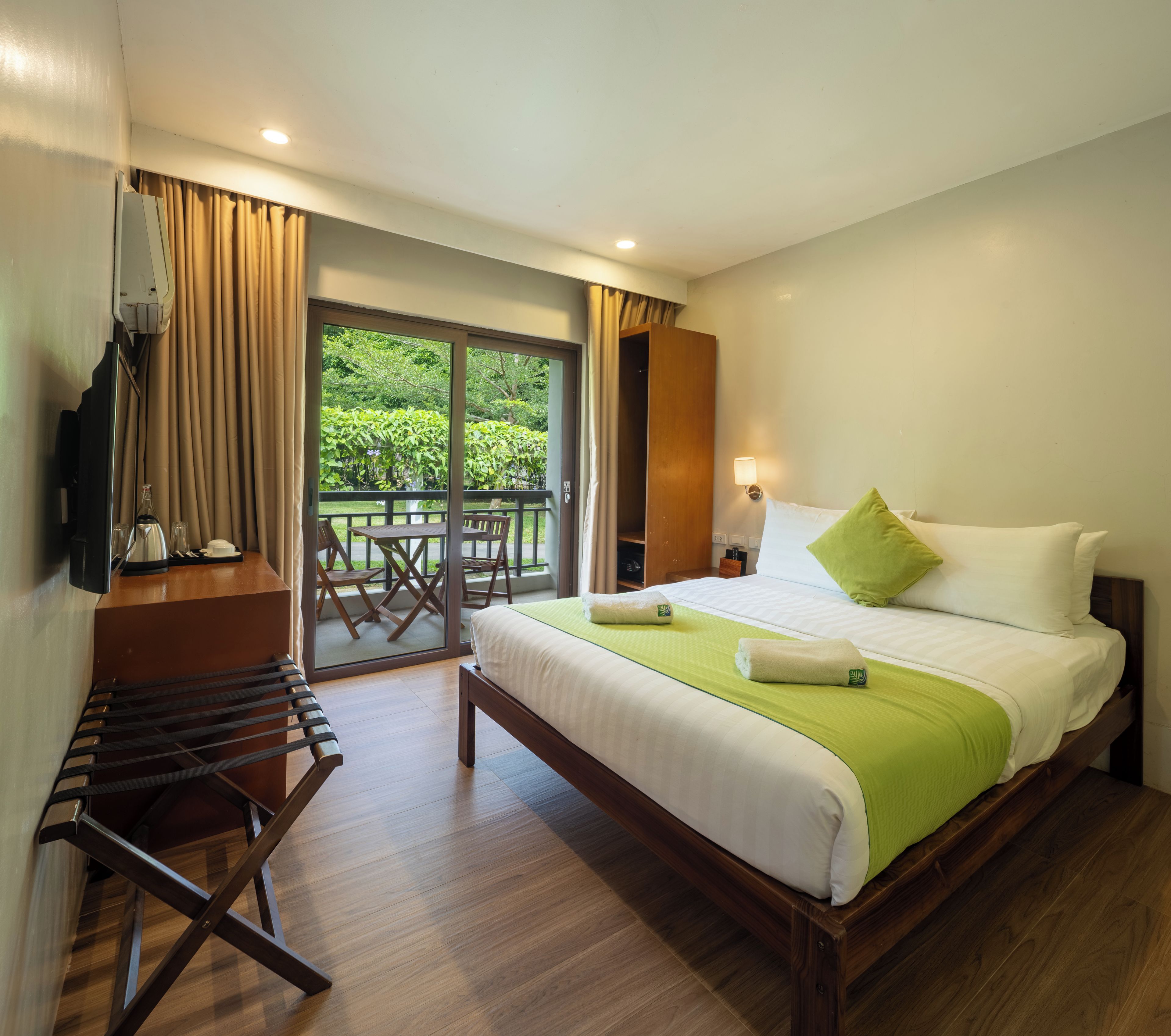 A hotel room with a bed, a TV, a table with chairs, and a balcony with a view of greenery.