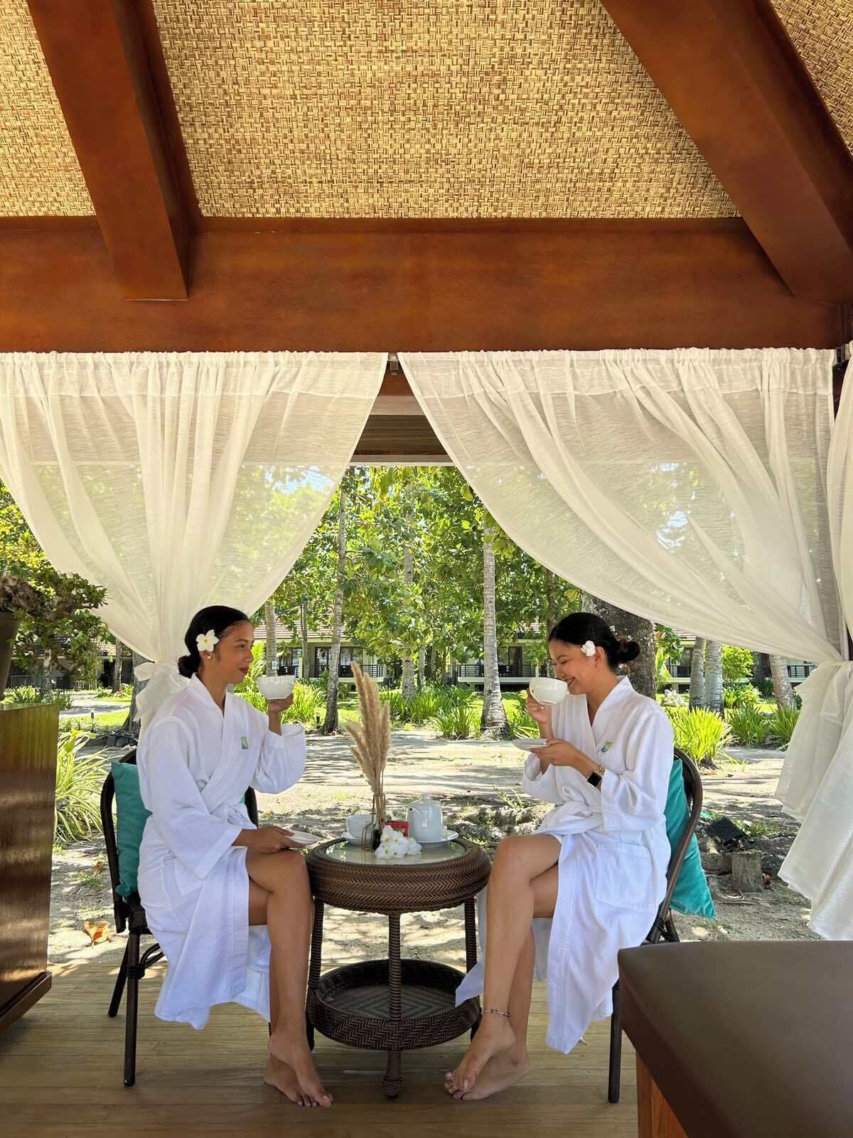 Two people in white robes sitting at a round table under a canopy with sheer curtains, with a tropical garden in the background.