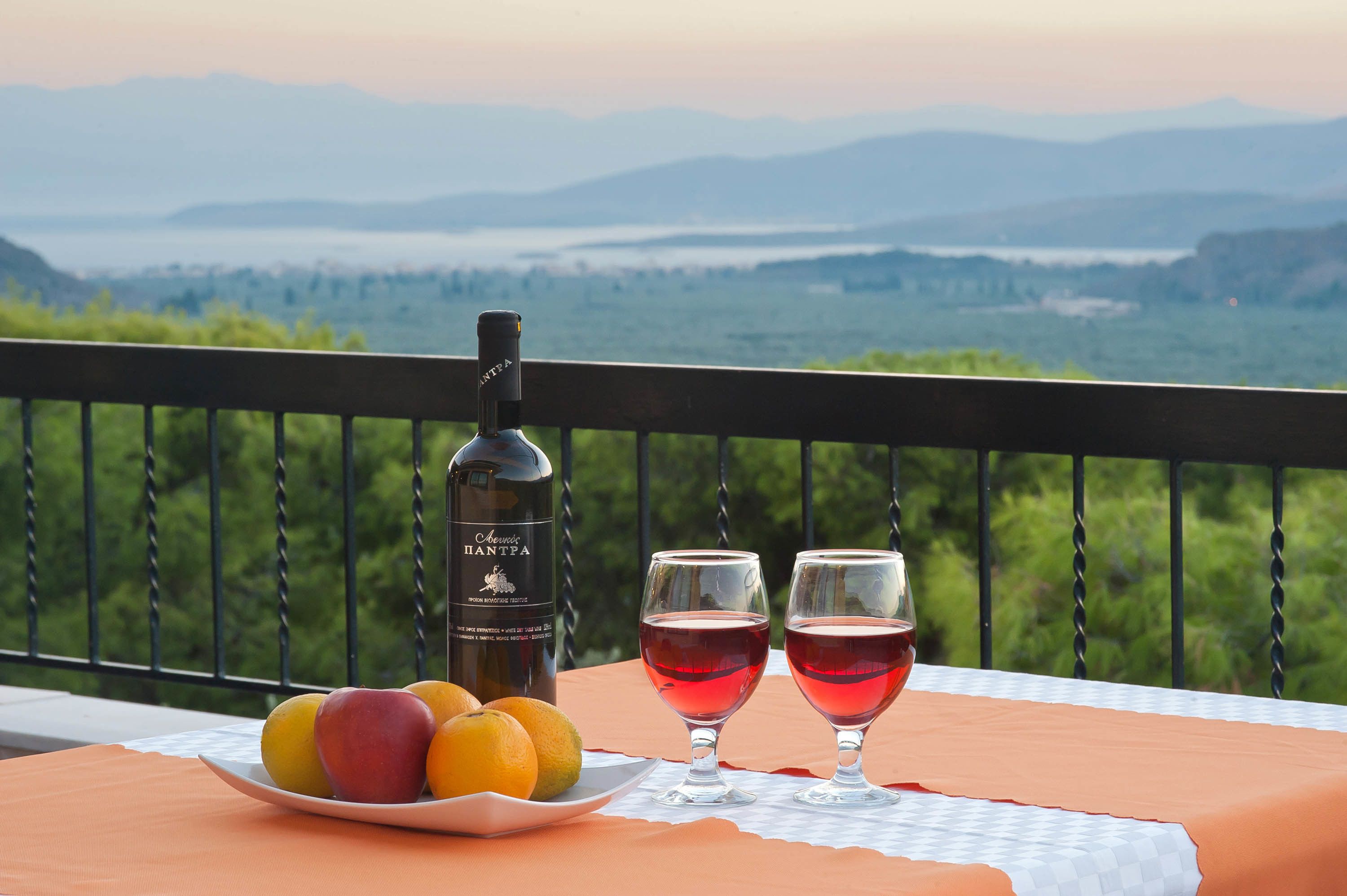 A table with a bottle of wine, two glasses of red wine, and a plate of fruit on a balcony overlooking a scenic landscape. - Chrissa Camping Rooms & Bungalows (Delphi)
