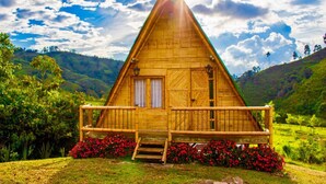 Une cabane triangulaire en bois, dotée d’un porche à l’avant, entourée d’une végétation luxuriante et de collines.