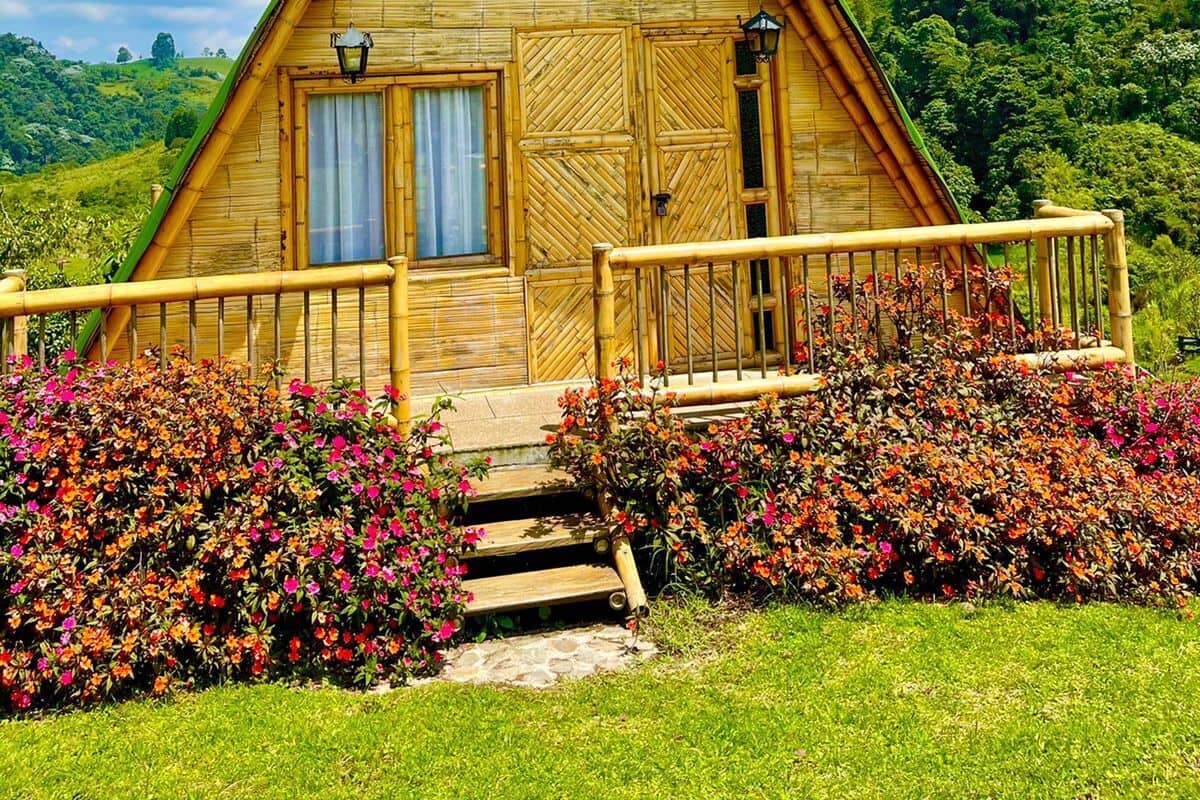 Een houten cabin in driehoekige vorm met een balkon en een trap die naar de ingang leidt, omringd door weelderige begroeiing en bloemen.