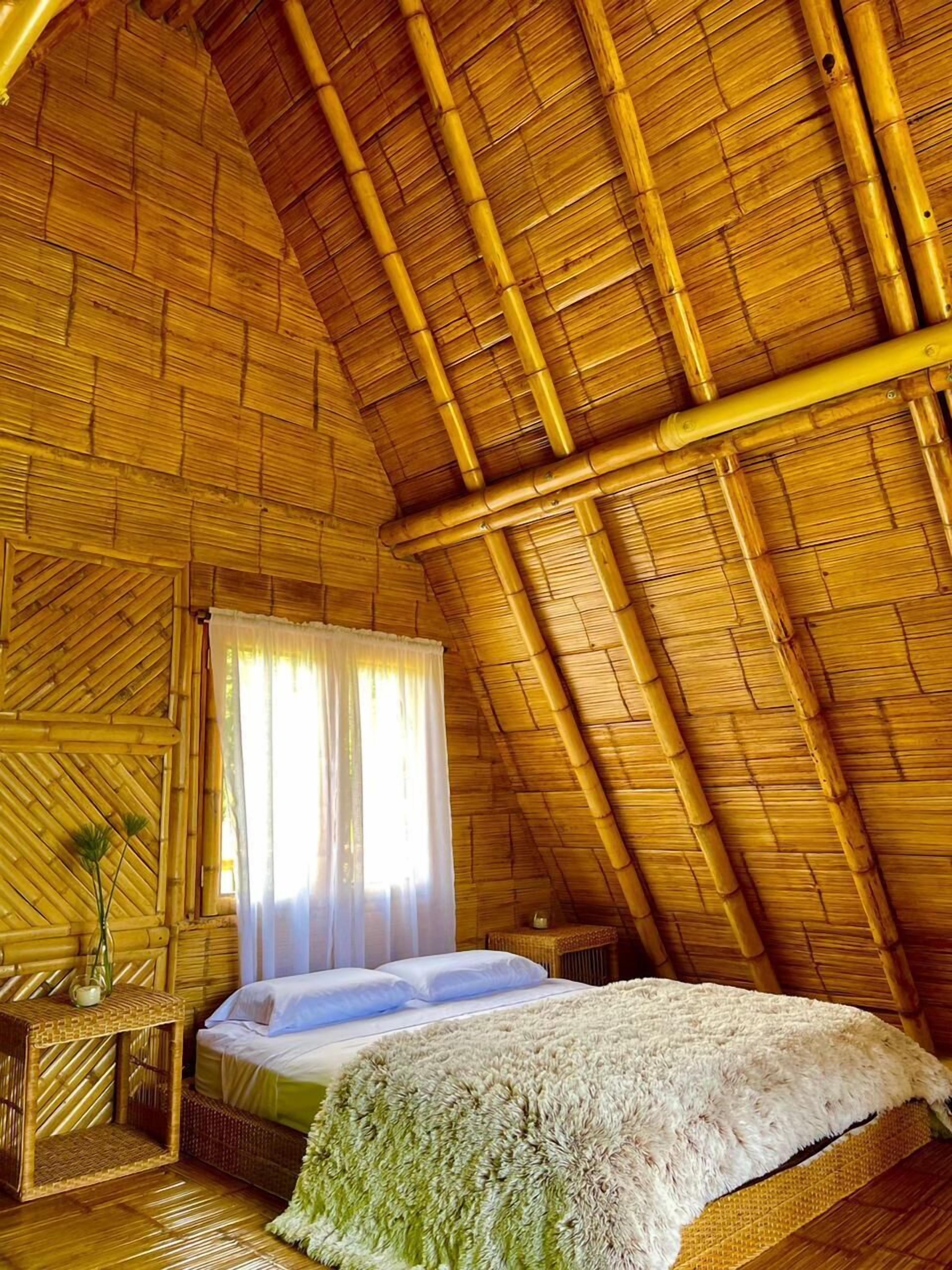 A room with a high, sloped bamboo ceiling, a bed with a white blanket, a small table with a vase, and a window with sheer curtains.