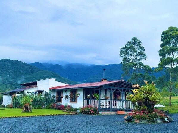 Casa de dos pisos con techo rojo, rodeada de abundante vegetación y vistas a montañas distantes.