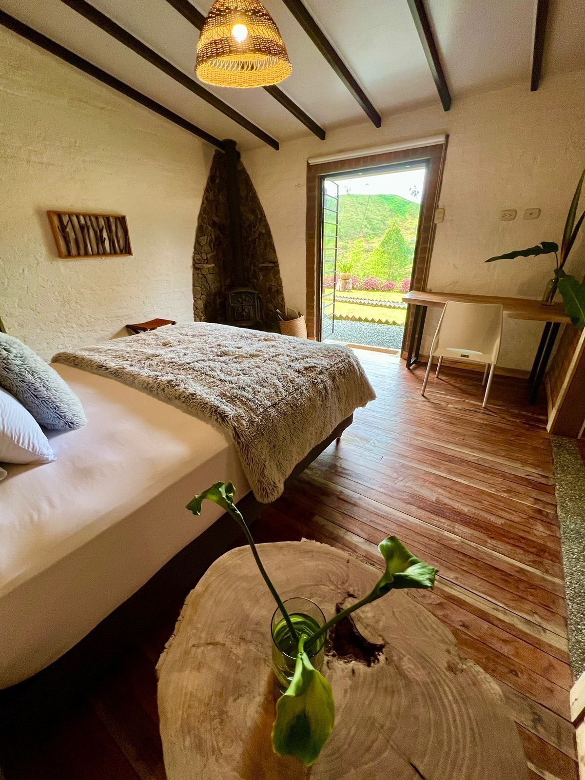 Een slaapkamer met een bed, een stenen open haard, een eethoek met een tafel en stoelen, en uitzicht op groen buiten.