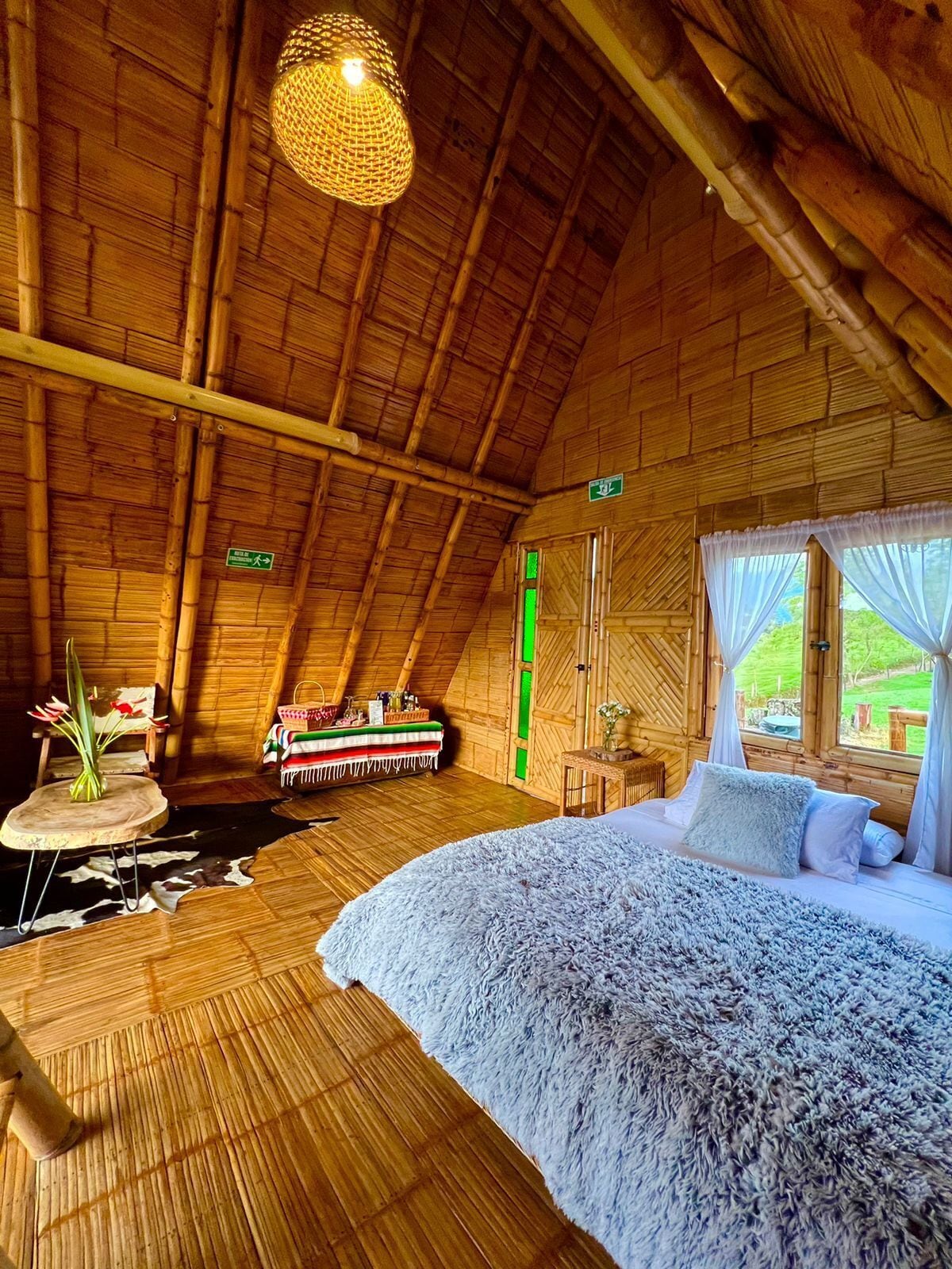 A cozy, wooden-roofed room with a bed, a hanging light fixture, and a view of greenery outside.