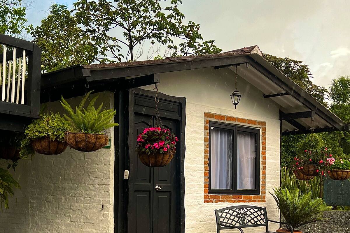 Een klein huis met een zwarte deur, een raam met gordijn, en een bankje ervoor. Er staan potplanten en een bloemenmand bij de deur.
