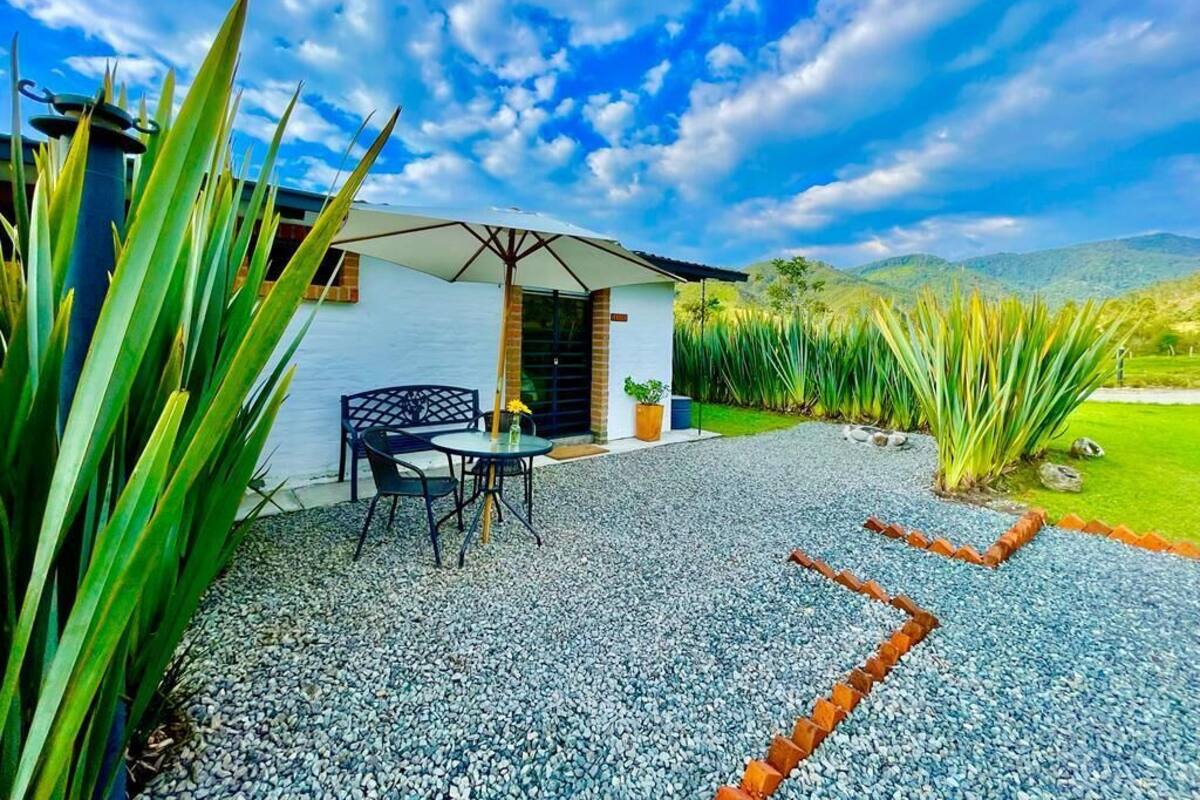 Een terras met een tafel en stoelen, een klein gebouw met een deur, en uitzicht op een grasveld en heuvels op de achtergrond.