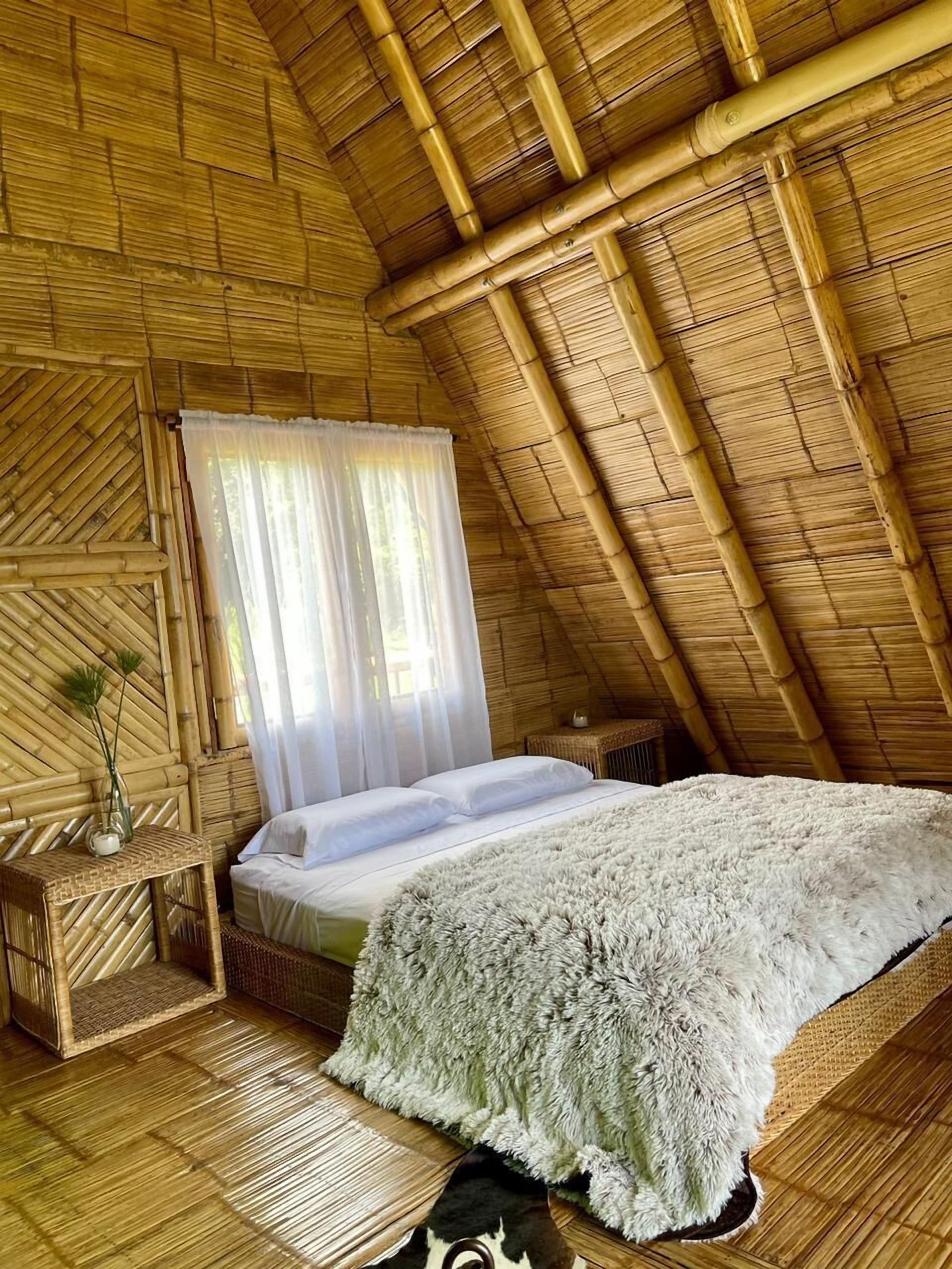 A room with a high, sloped ceiling made of bamboo, a bed with white linens, a small bedside table with a vase, and a woven rug on the floor.