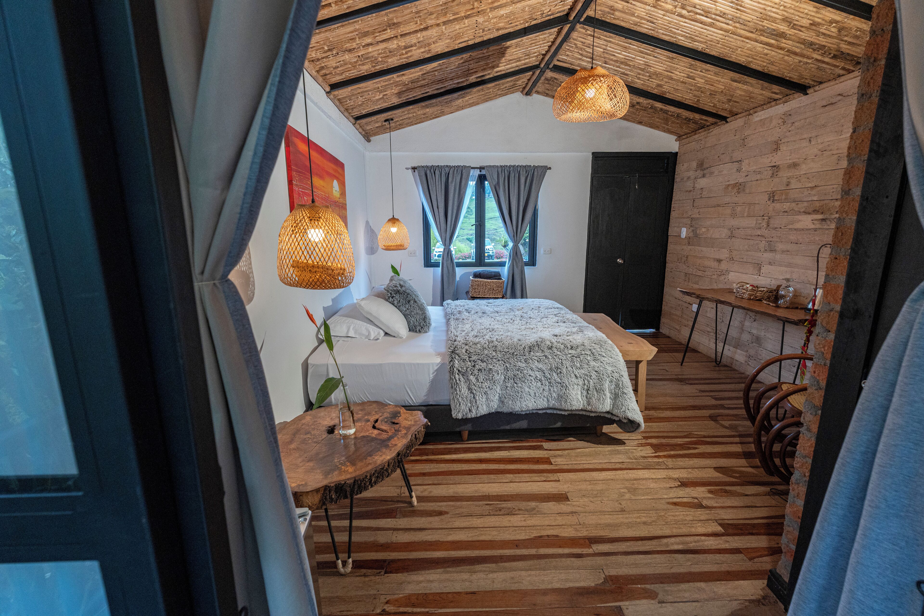 A cozy bedroom with a wooden ceiling, a bed with a gray blanket, a wooden table, and a hanging light fixture.