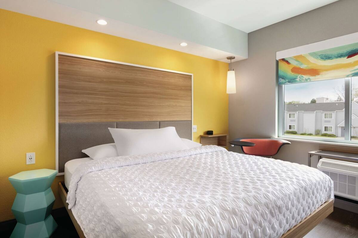 Ein modernes Hotelzimmer mit einem großen Bett, einem kleinen Nachttisch, einem Sessel und einem Fenster mit Blick auf die Stadt.
