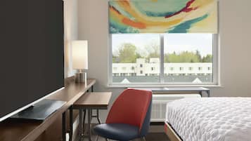A hotel room with a bed, a desk, a chair, a TV, and a window with a view of buildings and trees.