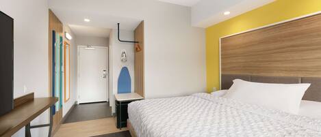 Habitación de hotel con cama, escritorio y espejo. Hay una puerta que conduce a un baño y un perchero.