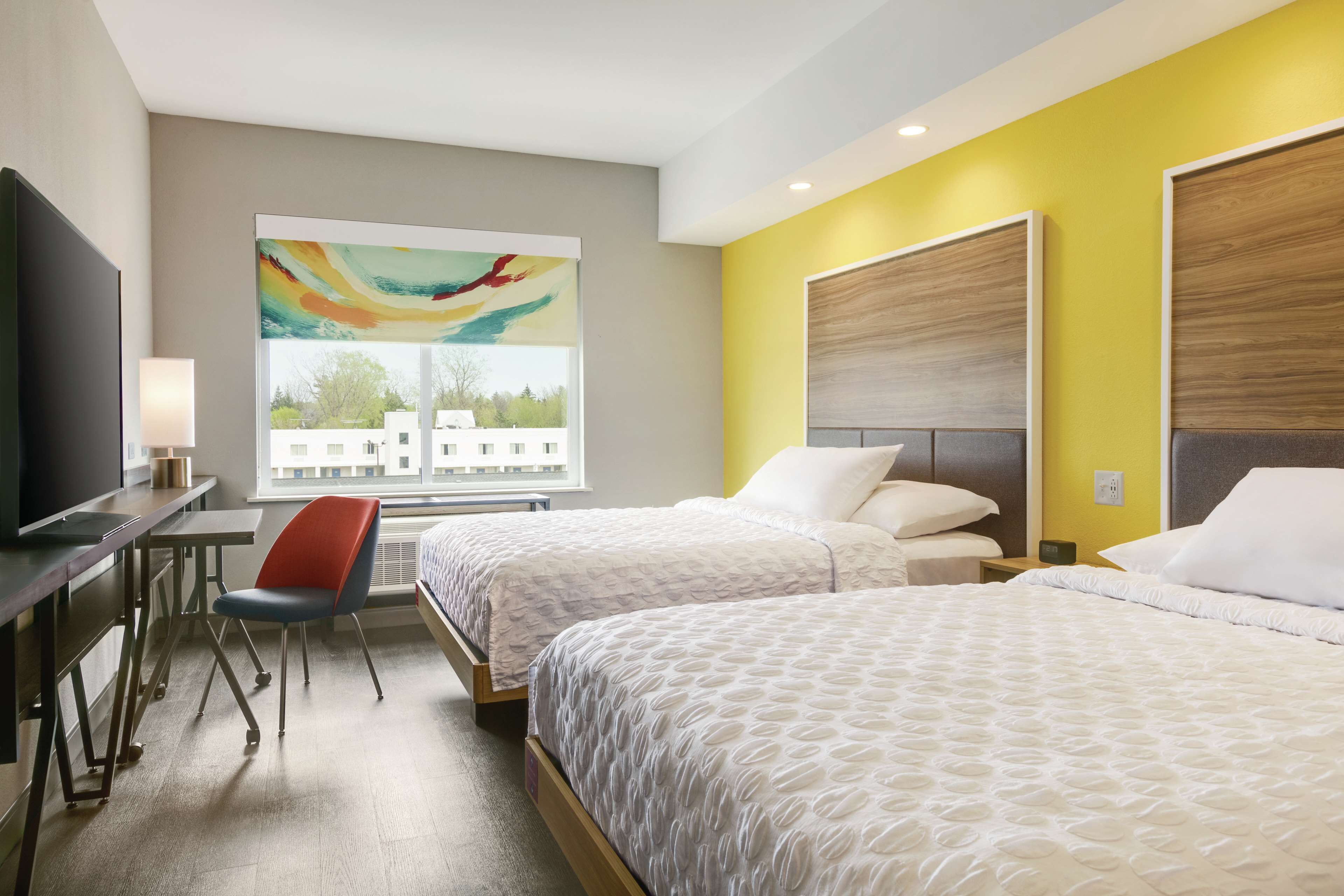 A hotel room with two beds, a desk with a chair, a TV, and a large window with a view of buildings.