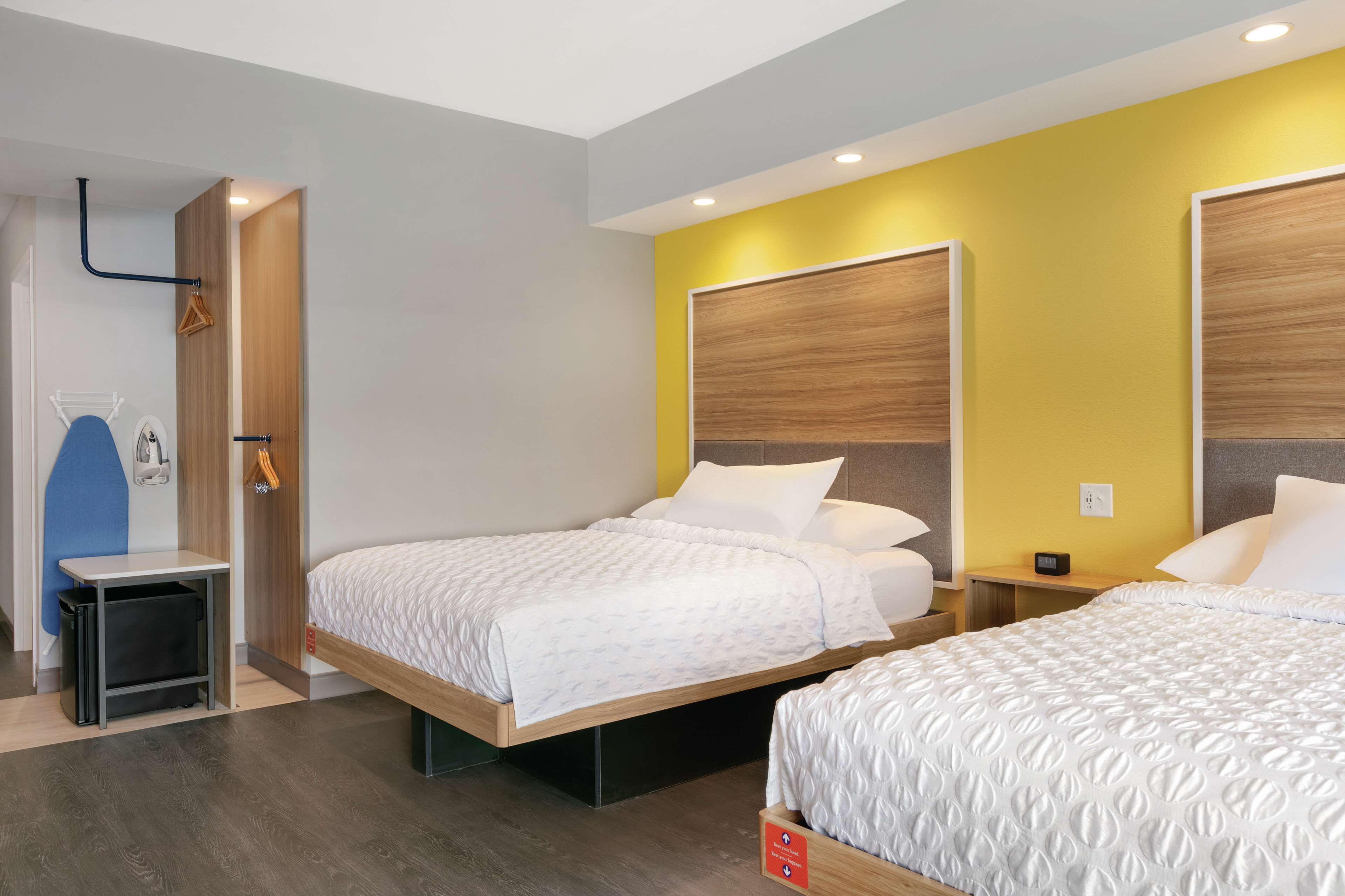 A hotel room with two beds, a wooden headboard, a small bedside table, and a wall-mounted towel rack.