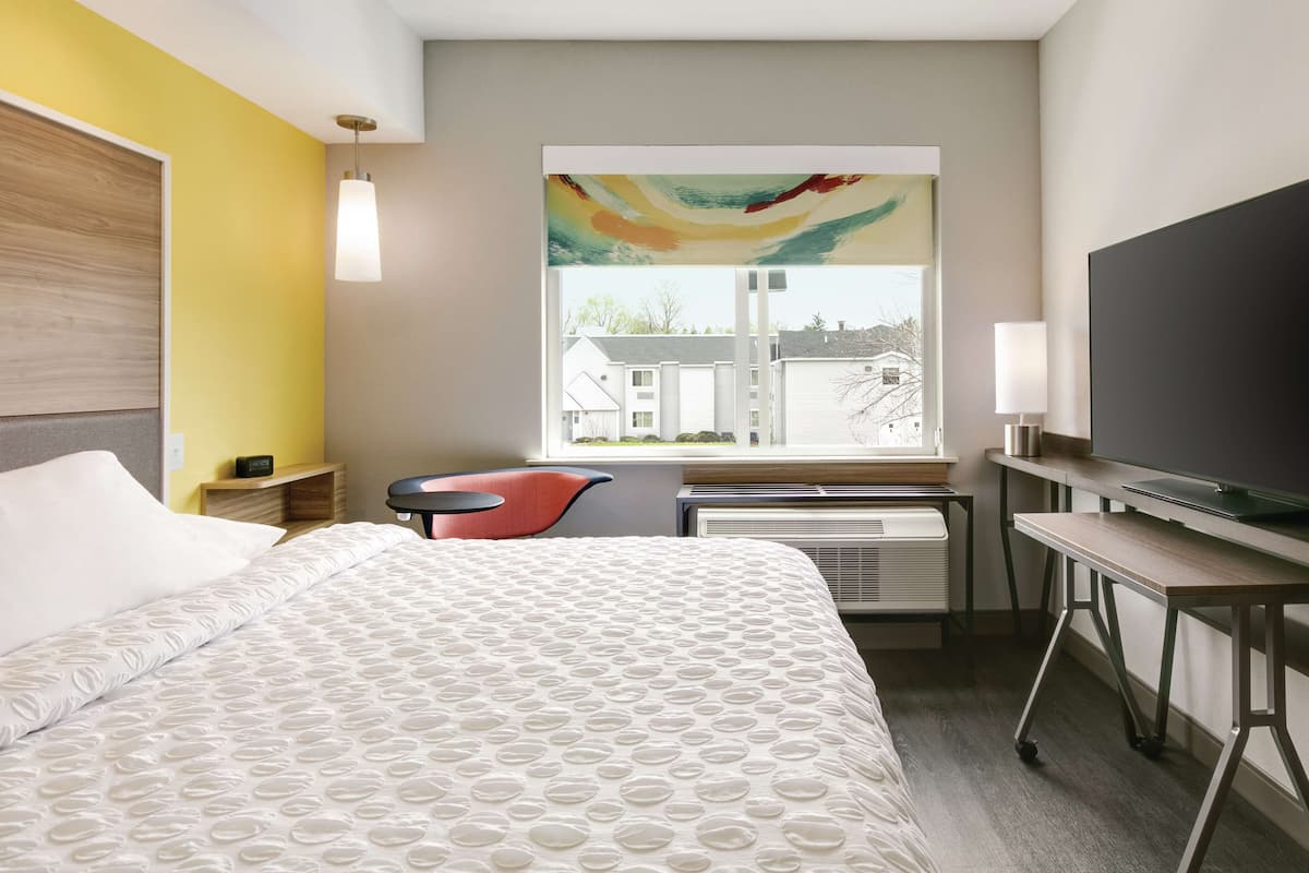 Une chambre d’hôtel équipée d’un lit, d’un bureau, d’une chaise, d’une télévision et d’une fenêtre donnant sur des immeubles.
