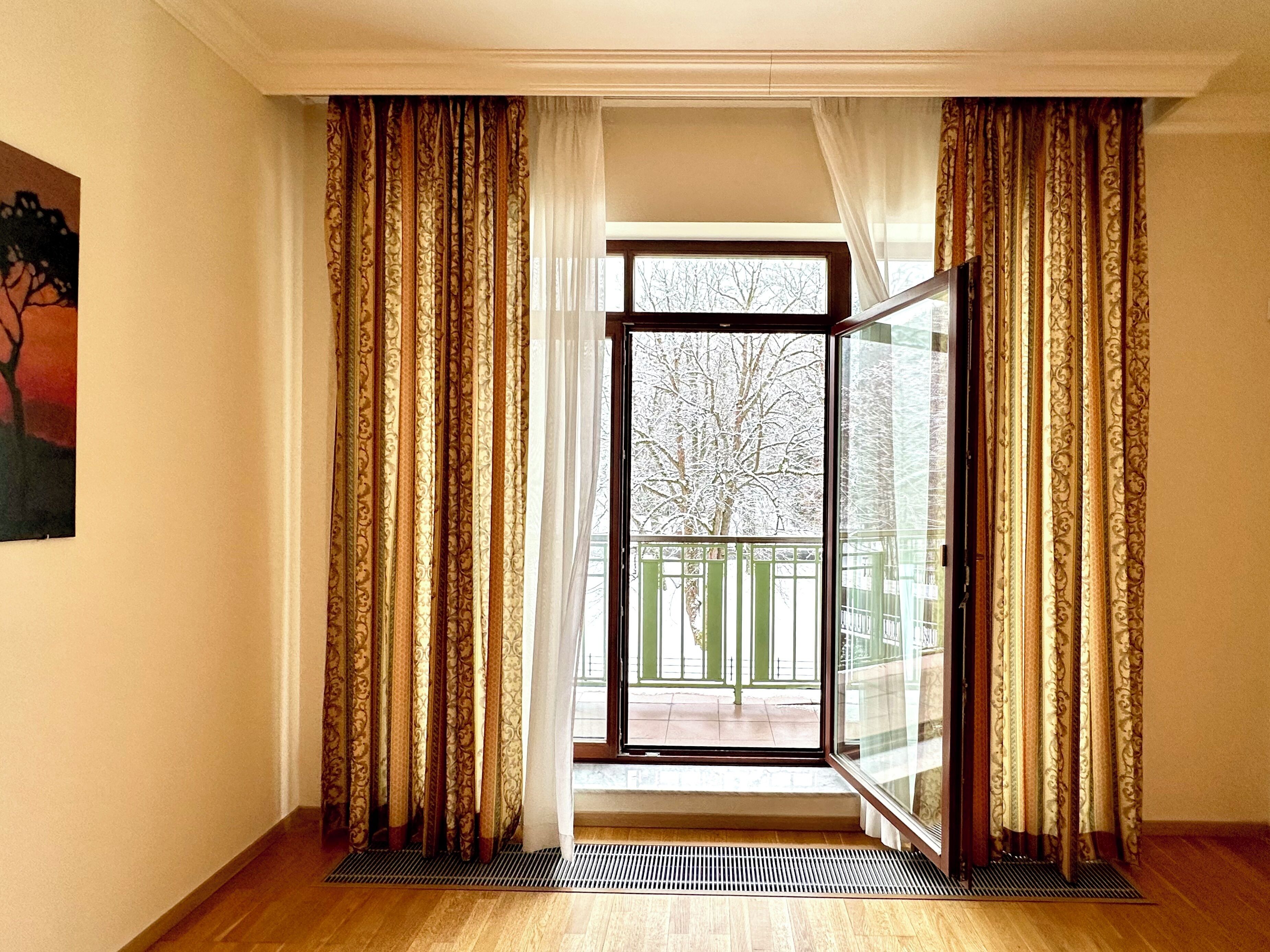 A room with a large sliding glass door, patterned curtains, a radiator, and a view of a balcony with a tree.